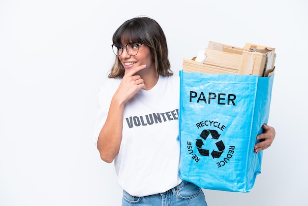 Mujer caucásica joven sosteniendo una bolsa de reciclaje llena de papel para reciclar aislado sobre fondo blanco pensando en una idea y mirando al lado