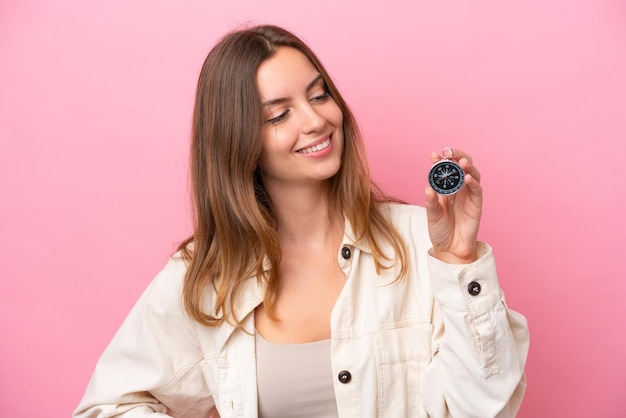 Mujer caucásica joven sosteniendo brújula aislada sobre fondo rosa con expresión feliz