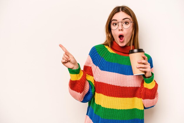 Mujer caucásica joven sosteniendo un café para llevar aislado sobre fondo blanco apuntando hacia un lado