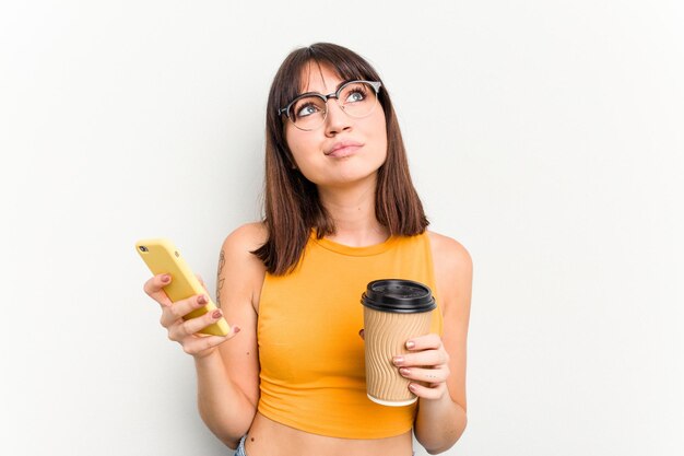 Mujer caucásica joven sosteniendo un café para llevar y un teléfono móvil aislado sobre fondo blanco.