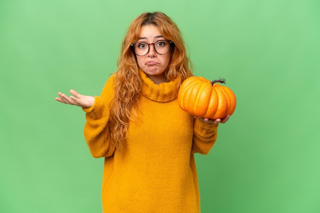 Mujer caucásica joven sosteniendo una calabaza aislada en el fondo de la clave de croma de pantalla verde que tiene dudas mientras levanta las manos