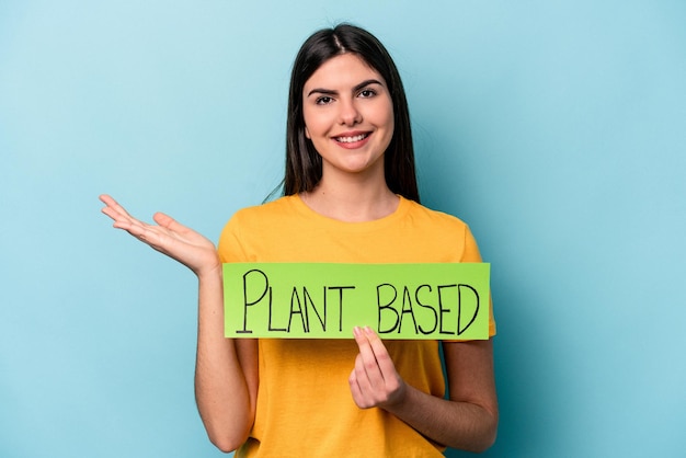 Mujer caucásica joven sosteniendo un cartel basado en plantas aislado en un fondo azul que muestra un espacio de copia en una palma y sosteniendo otra mano en la cintura