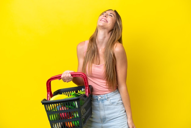 Mujer caucásica joven sosteniendo una cesta de la compra llena de comida aislada sobre fondo amarillo riendo