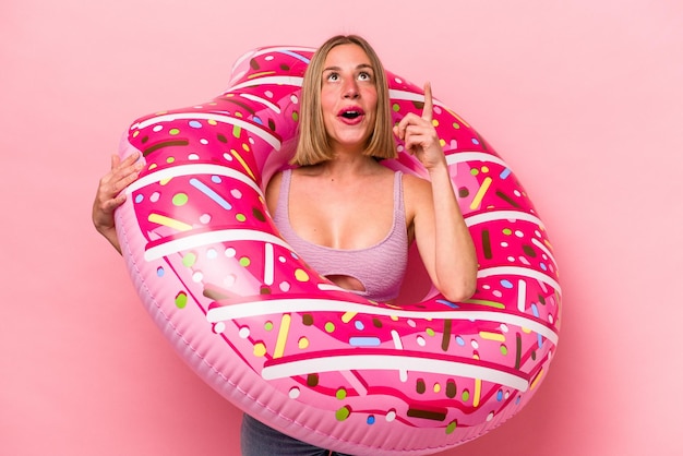 Mujer caucásica joven sosteniendo un colchón de aire aislado sobre fondo rosa apuntando hacia arriba con la boca abierta