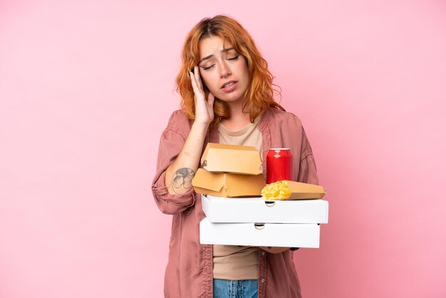 Foto mujer caucásica joven sosteniendo comida rápida aislada sobre fondo rosa con dolor de cabeza