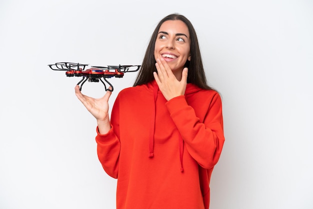 Mujer caucásica joven sosteniendo un dron aislado sobre fondo blanco mirando hacia arriba mientras sonríe