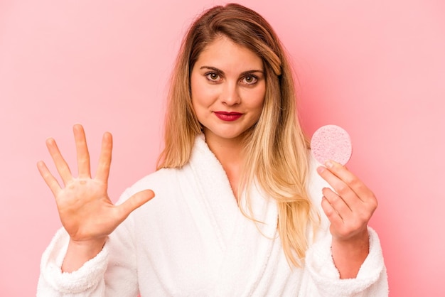 Mujer caucásica joven sosteniendo esponja facial aislada sobre fondo rosa sonriendo alegre mostrando el número cinco con los dedos