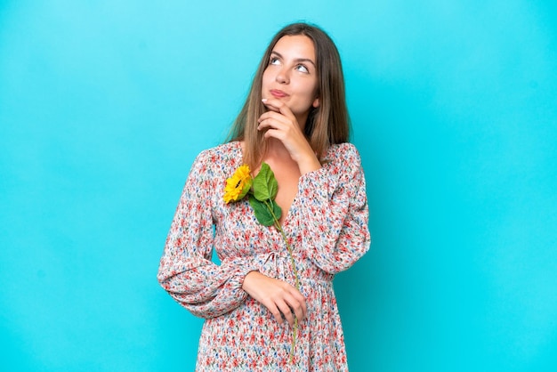 Mujer caucásica joven sosteniendo girasol aislado sobre fondo azul y mirando hacia arriba