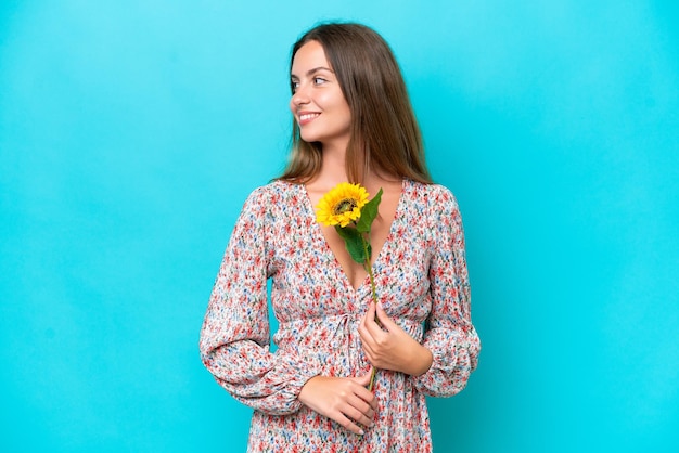 Mujer caucásica joven sosteniendo girasol aislado sobre fondo azul mirando hacia el lado