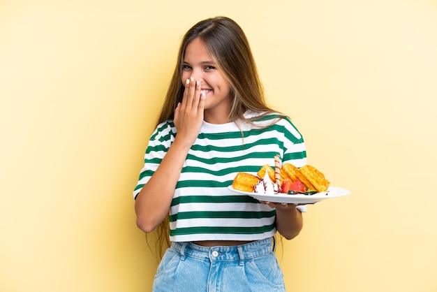 Mujer caucásica joven sosteniendo gofres aislados sobre fondo amarillo feliz y sonriente cubriendo la boca con la mano