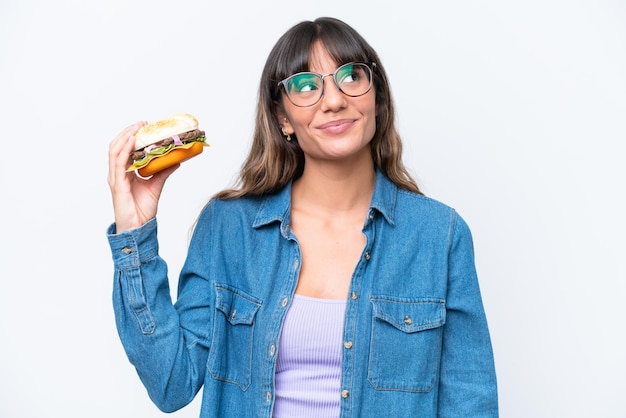 Mujer caucásica joven sosteniendo una hamburguesa aislada de fondo blanco mirando hacia arriba mientras sonríe