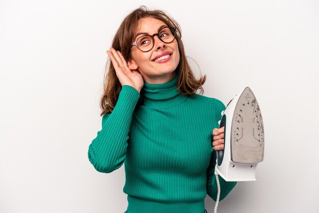 Mujer caucásica joven sosteniendo hierro aislado sobre fondo blanco tratando de escuchar un chisme