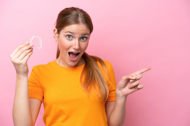 Mujer caucásica joven sosteniendo invising aislada sobre fondo rosa sorprendida y señalando con el dedo hacia un lado