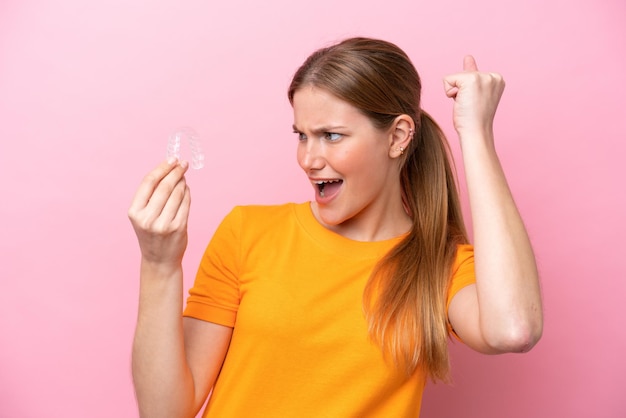 Mujer caucásica joven sosteniendo invising aislado sobre fondo rosa celebrando una victoria