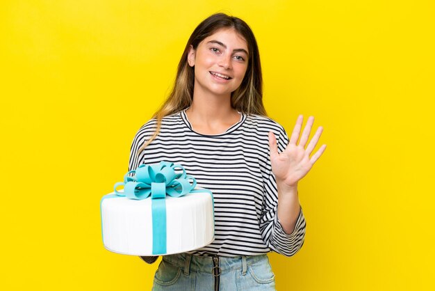 Foto mujer caucásica joven sosteniendo pastel de cumpleaños aislado sobre fondo amarillo contando cinco con los dedos