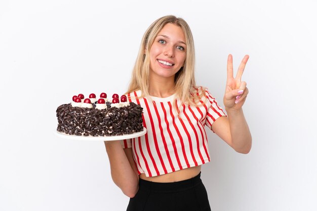 Mujer caucásica joven sosteniendo pastel de cumpleaños aislado sobre fondo blanco sonriendo y mostrando el signo de la victoria