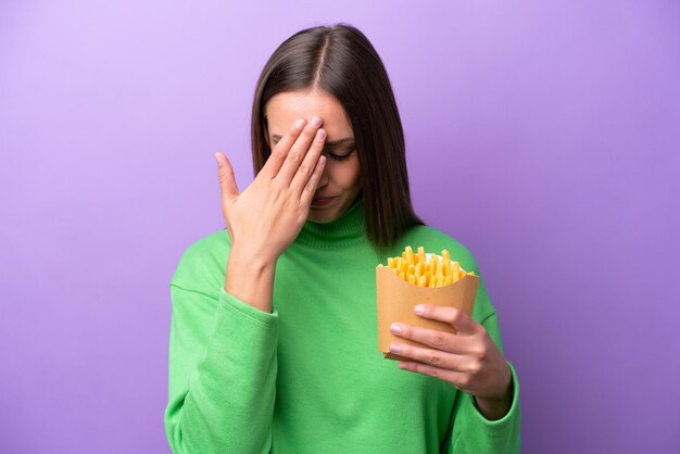 Mujer caucásica joven sosteniendo patatas fritas sobre fondo morado con expresión cansada y enferma