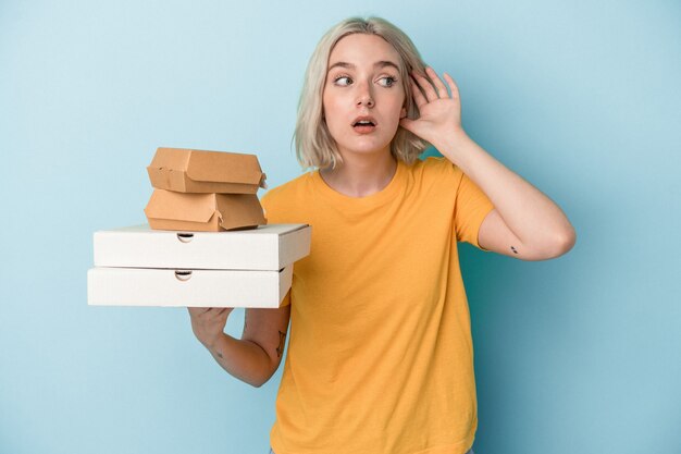 Mujer caucásica joven sosteniendo pizzas y hamburguesas aisladas sobre fondo azul tratando de escuchar un chisme.