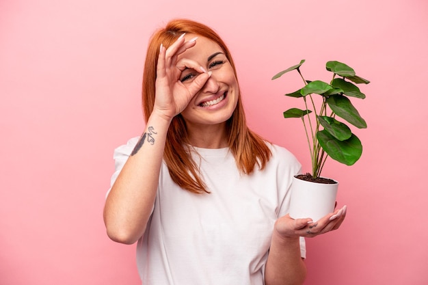 Mujer caucásica joven sosteniendo una planta aislada sobre fondo rosa Mujer caucásica joven sosteniendo una planta aislada sobre fondo rosa emocionada manteniendo el gesto correcto en el ojo.