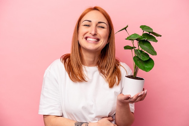 Mujer caucásica joven sosteniendo una planta aislada sobre fondo rosa Mujer caucásica joven sosteniendo una planta aislada sobre fondo rosa riendo y divirtiéndose.