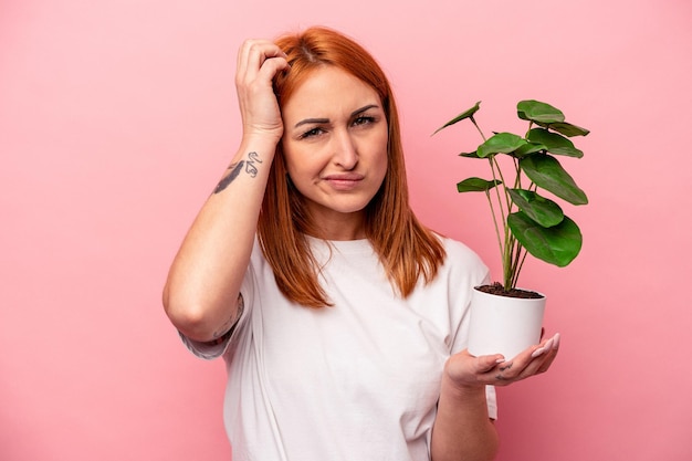 Mujer caucásica joven sosteniendo una planta aislada sobre fondo rosa Mujer caucásica joven sosteniendo una planta aislada sobre fondo rosa sorprendida, ha recordado una reunión importante.