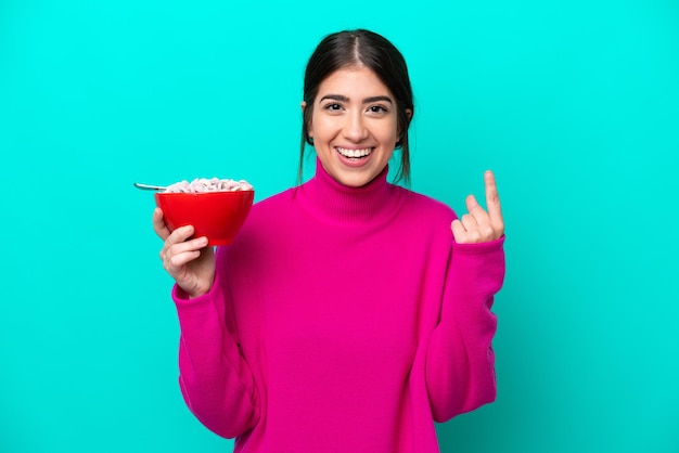 Mujer caucásica joven sosteniendo un plato de cereales aislado sobre fondo azul apuntando a una gran idea