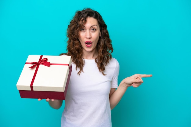 Mujer caucásica joven sosteniendo un regalo aislado sobre fondo azul sorprendido y apuntando hacia el lado