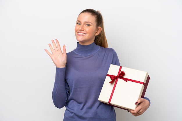 Mujer caucásica joven sosteniendo un regalo aislado sobre fondo blanco saludando con la mano con expresión feliz