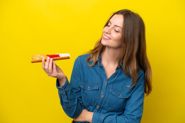 Mujer caucásica joven sosteniendo sashimi aislado sobre fondo amarillo con expresión feliz