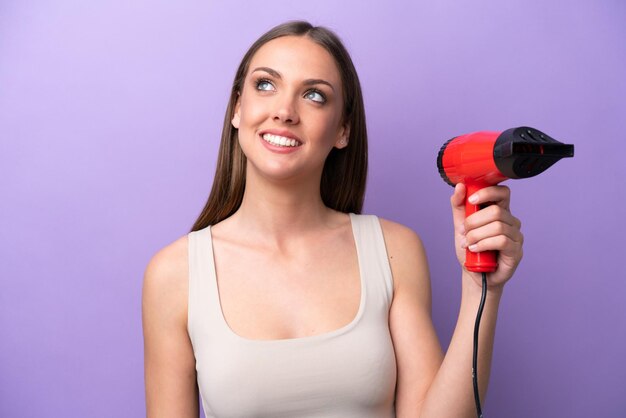 Mujer caucásica joven sosteniendo un secador de pelo aislado en un fondo morado mirando hacia arriba mientras sonríe