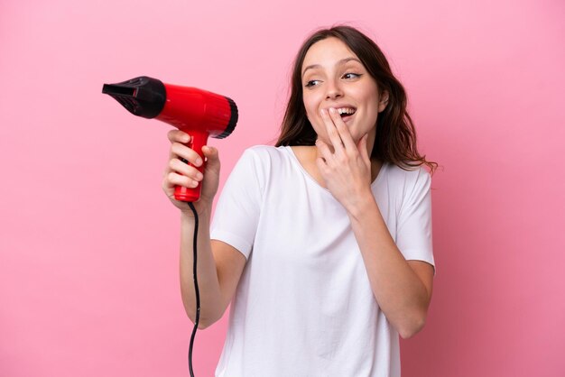 Mujer caucásica joven sosteniendo un secador de pelo aislado en un fondo rosa con expresión facial sorprendida y sorprendida