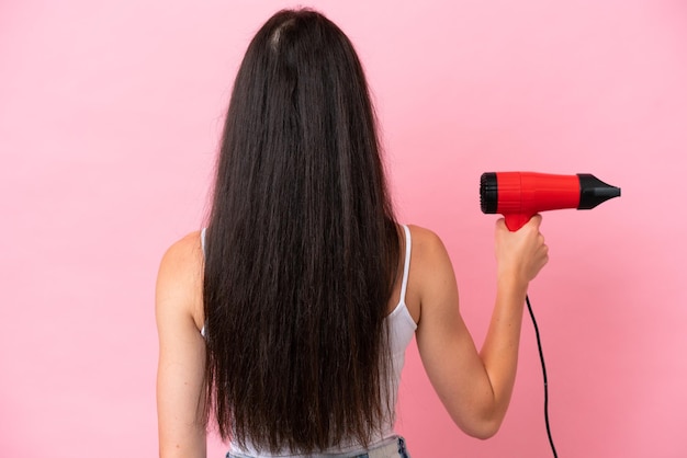 Mujer caucásica joven sosteniendo un secador de pelo aislado sobre fondo rosa en posición trasera