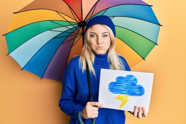 Mujer caucásica joven sosteniendo un sorteo de lluvia y un paraguas inflando las mejillas con la cara graciosa boca inflada con aire atrapando aire