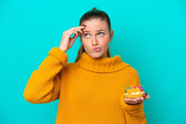 Mujer caucásica joven sosteniendo una tartaleta aislada de fondo azul que tiene dudas y con una expresión facial confusa