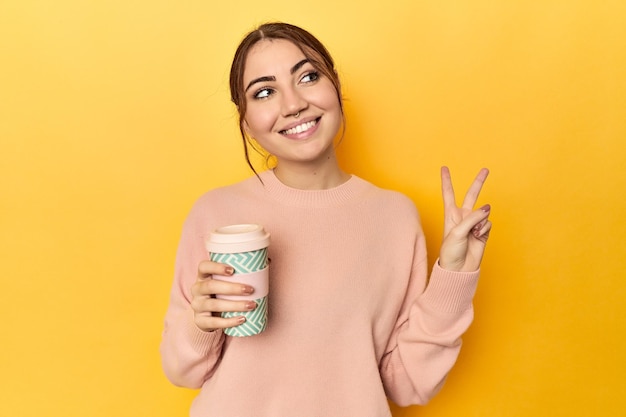 Mujer caucásica joven sosteniendo una taza de café para llevar alegre y despreocupada mostrando un símbolo de paz