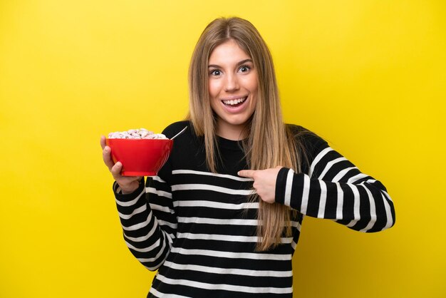 Mujer caucásica joven sosteniendo un tazón de cereales aislado sobre fondo amarillo con expresión facial sorpresa