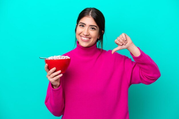 Mujer caucásica joven sosteniendo un tazón de cereales aislado sobre fondo azul orgullosa y satisfecha