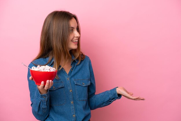 Mujer caucásica joven sosteniendo un tazón de cereales aislado sobre fondo rosa con expresión facial sorpresa