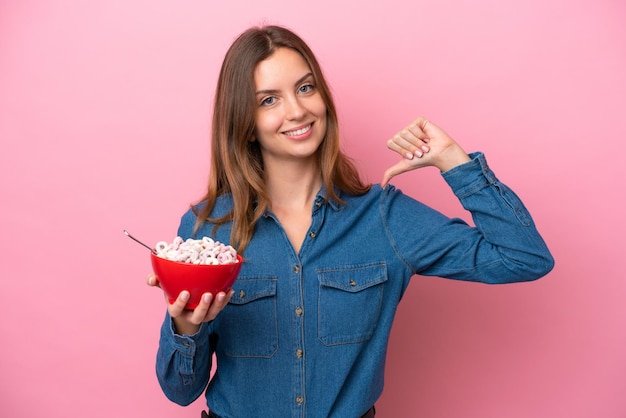 Mujer caucásica joven sosteniendo un tazón de cereales aislado sobre fondo rosa orgullosa y satisfecha