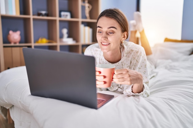 Mujer caucásica joven usando laptop bebiendo café en el dormitorio