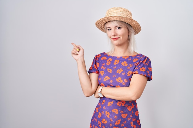 Mujer caucásica joven con vestido de flores y sombrero de verano apuntando con el dedo de la mano hacia un lado mostrando publicidad cara seria y tranquila