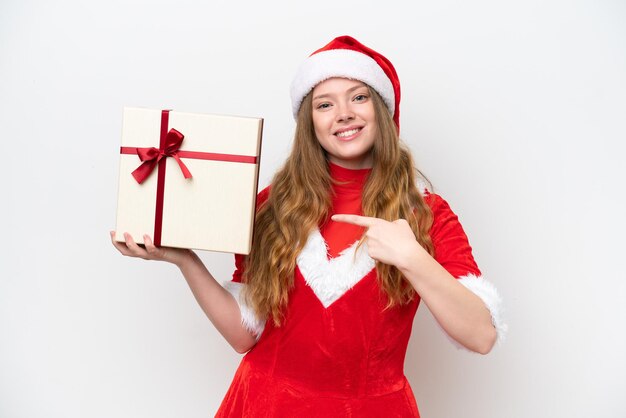 Mujer caucásica joven con vestido de Navidad sosteniendo regalo aislado sobre fondo blanco y apuntándolo