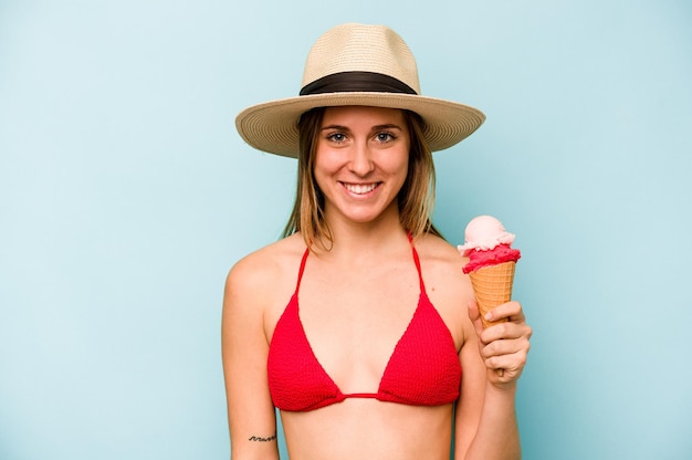 Mujer caucásica joven vistiendo un bikini y sosteniendo un helado aislado sobre fondo azul feliz sonriendo y alegre