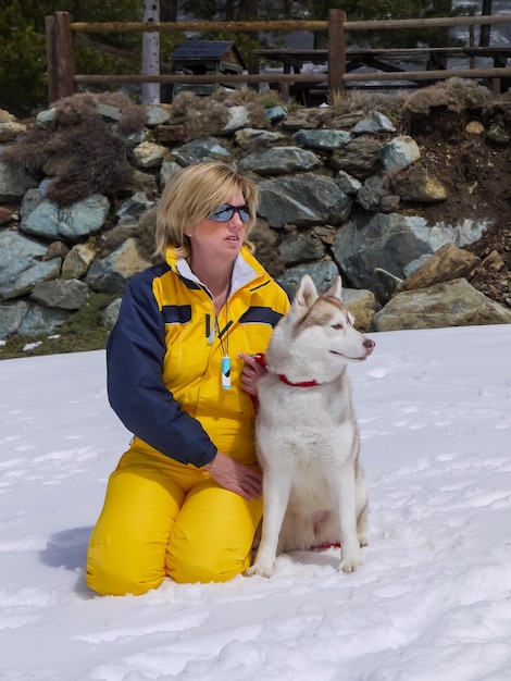 Mujer caucásica madura rubia con cristal de nieve arrodillada cerca del perro husky siberiano en la nieve