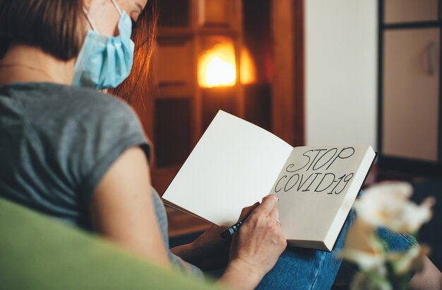 Mujer caucásica con una máscara especial está escribiendo algo en un libro mientras está sentado en el sofá