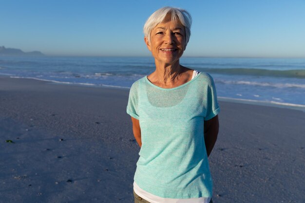 Mujer caucásica mayor disfrutando del tiempo en la playa en un día soleado, mirando a la cámara y sonriendo con el cielo azul y el mar de fondo