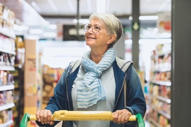 Foto mujer caucásica mayor empujando un carrito de compras en el supermercado mirando los productos en exhibición al cliente atento a los costos de compra