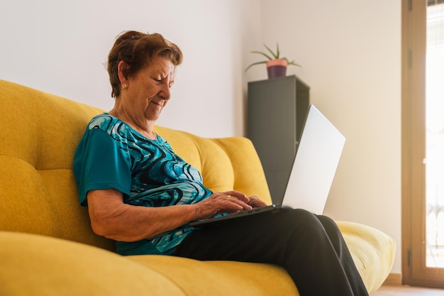 Foto mujer caucásica mayor escribiendo en una laptop sentada en un sofá amarillo lleva una camiseta azul natura