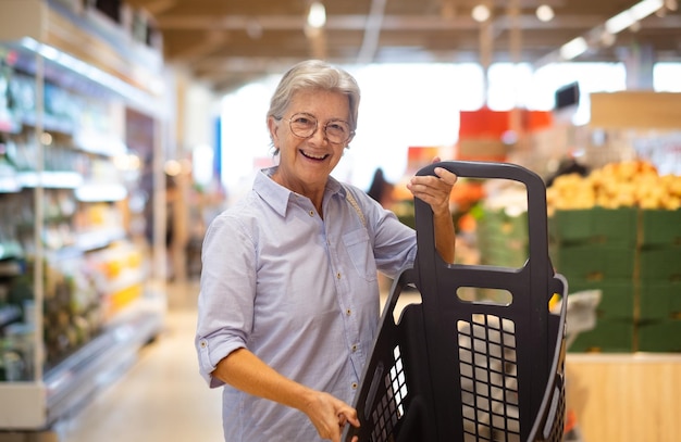 Mujer caucásica mayor en el supermercado sosteniendo un carrito de compras mirando el aumento de precios de la cámara y la inflación