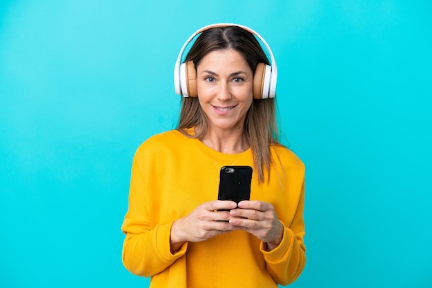 Mujer caucásica de mediana edad aislada de fondo azul escuchando música con un móvil y mirando al frente